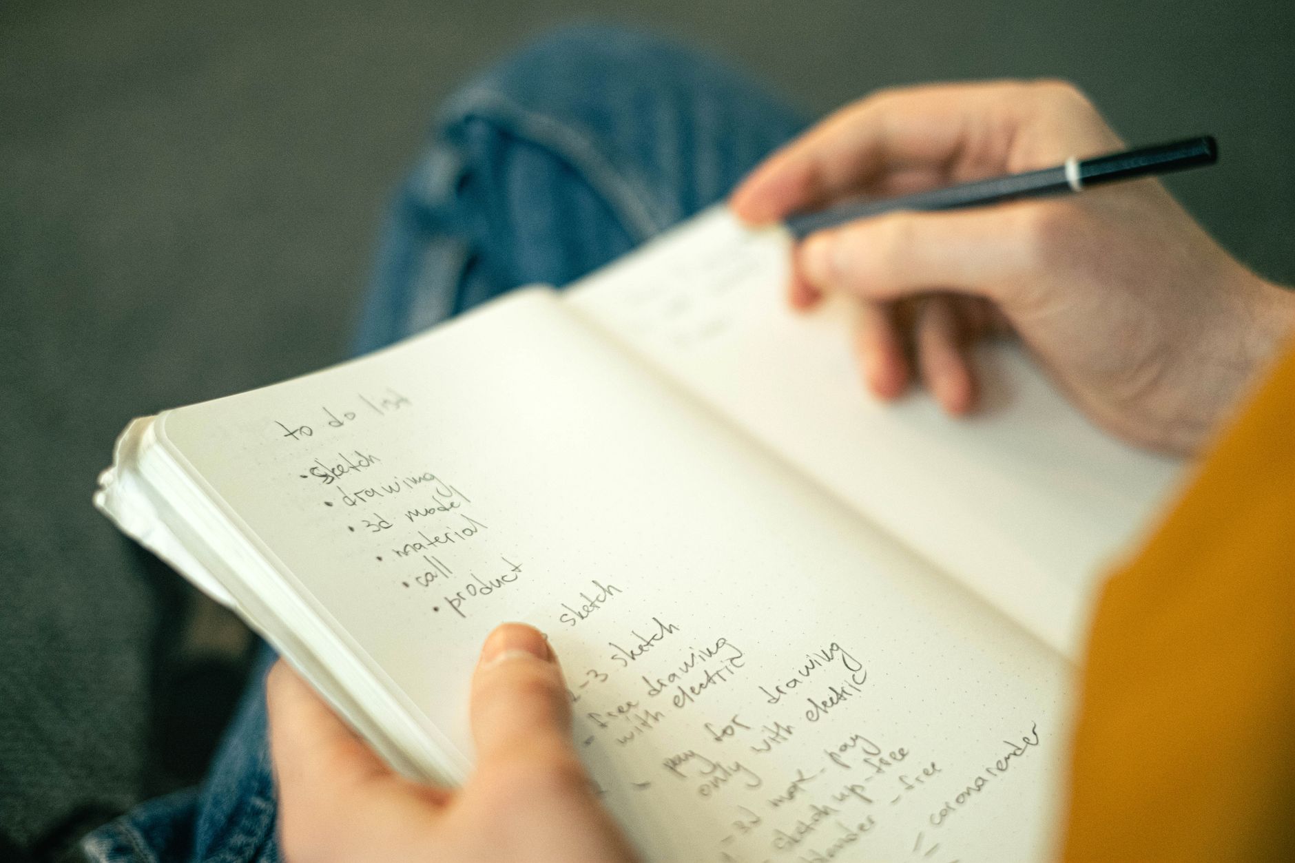 膝の上で開いたノートに、ペンで箇条書きのやることリストを書き込んでいる手元。いくつかの項目にチェックが入っている。