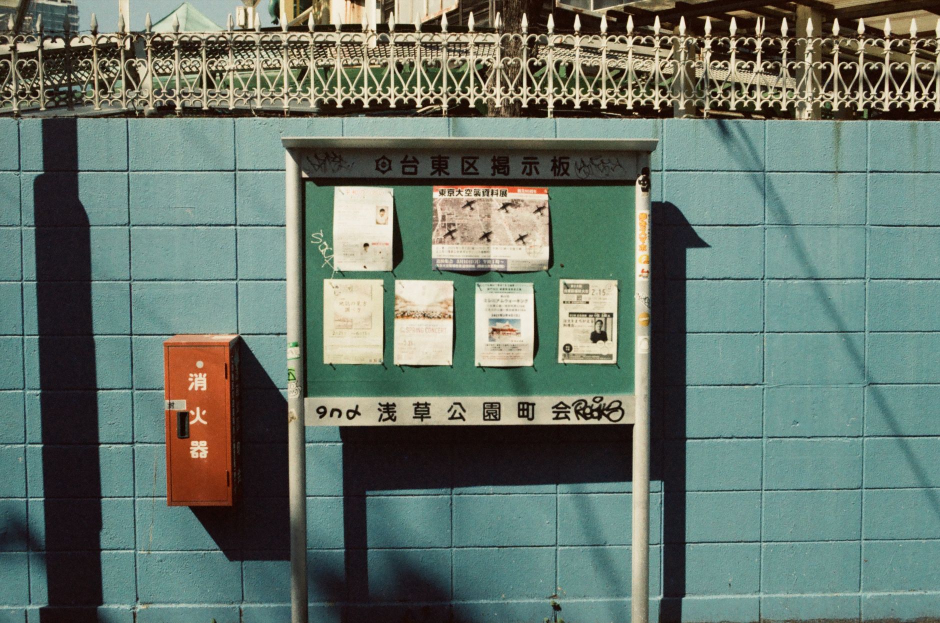 水色のブロック塀に取り付けられた町内会の掲示板に、白い紙のお知らせが数枚ならべて貼られている。